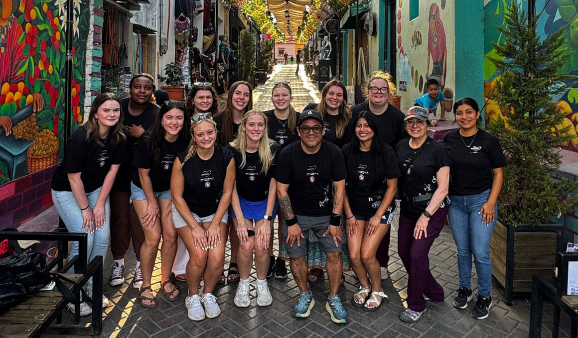 A group of about a dozen USD students are wearing matching black t-shirts, posing together in a brightly painted alley in Guatemala, smiling at the camera with murals and hanging decorations behind them.