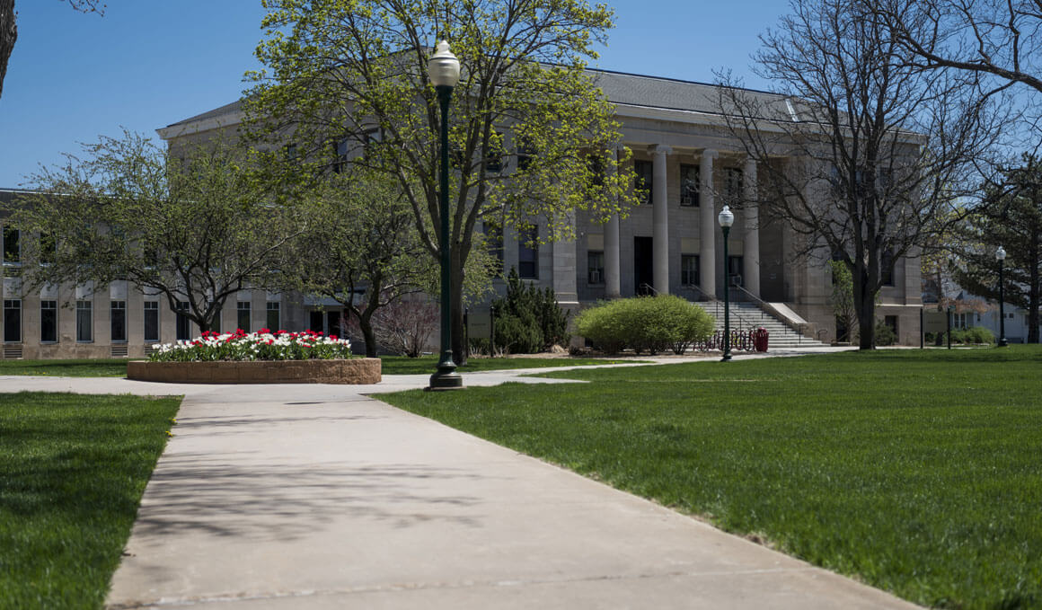 This photo shows the exterior of the College of Arts & Sciences, surrounded by blue sky, green grass, trees and flowers.