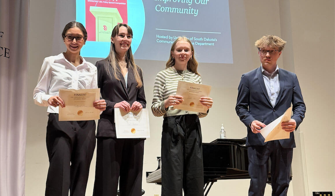 USD students stand on a stage, in front of a screen, holding their certificates for placing in the Speakers' Night competition.