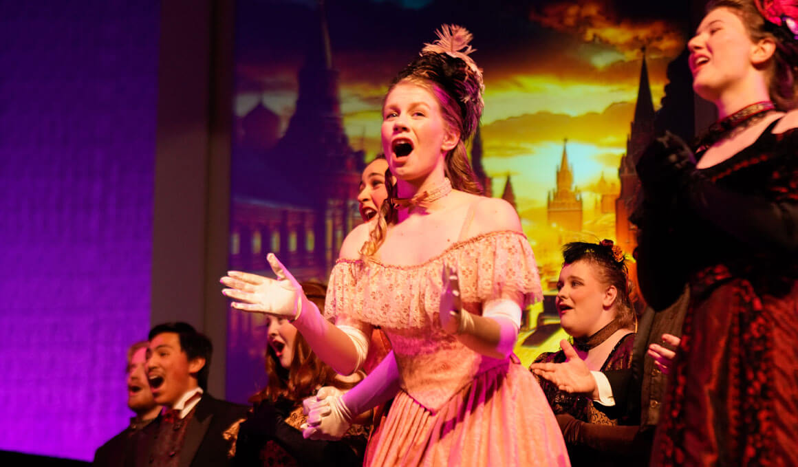 USD Opera students stand on stage during a performance. They are in hats, dresses, and suits of various colors. In the background there is a backdrop with castles and clouds on it.