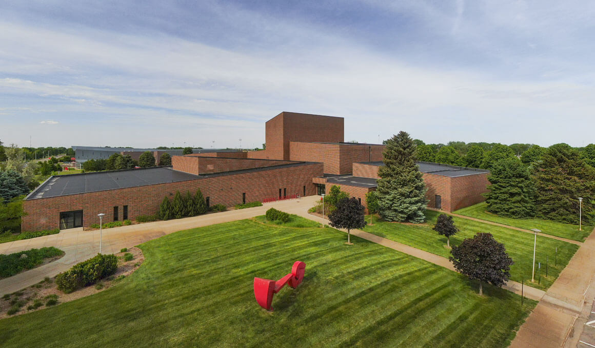 An exterior drone photo of the Warren M. Lee Center for Fine Arts on the USD campus. It is surrounded by green lawns, green trees and a blue sky. One of the lawns has a red statue on it.