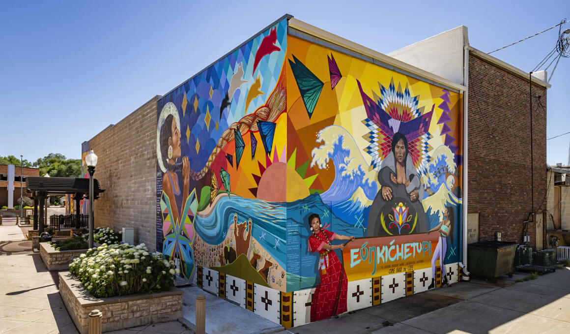 A vibrant, multi-colored outdoor mural depicting figures and cultural symbols wraps around the corner of a brick building in downtown Vermillion.