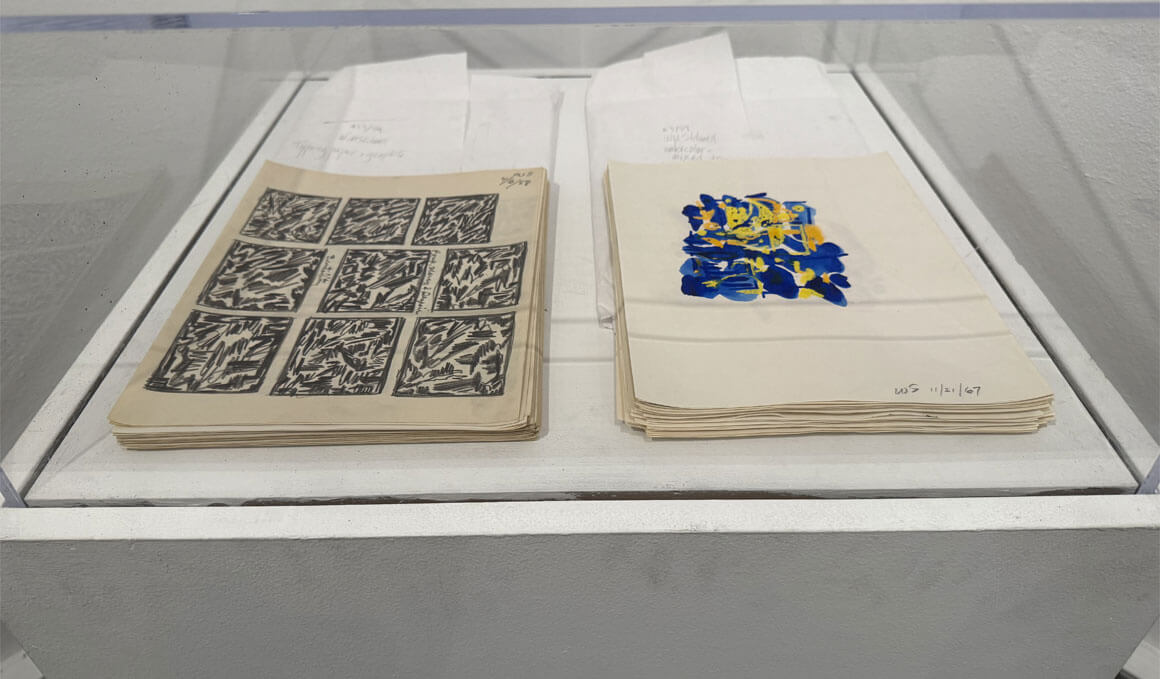 Two stacks of cream-colored paper artworks, by Wilber Stilwell, sit side by side in a glass display case, with black sketch grid drawings on the left and a blue-and-yellow abstract paint on the right.