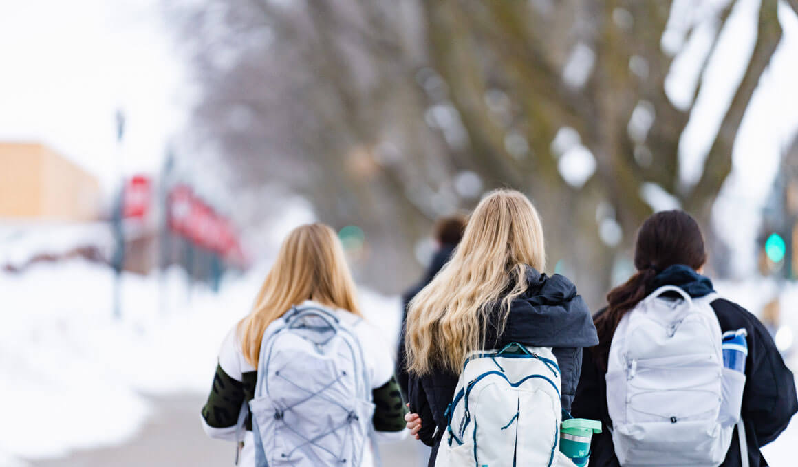 Three USD students walk on the snowy campus, with their backs turned to the camera. They are wearing their backpacks, with coats and hats.