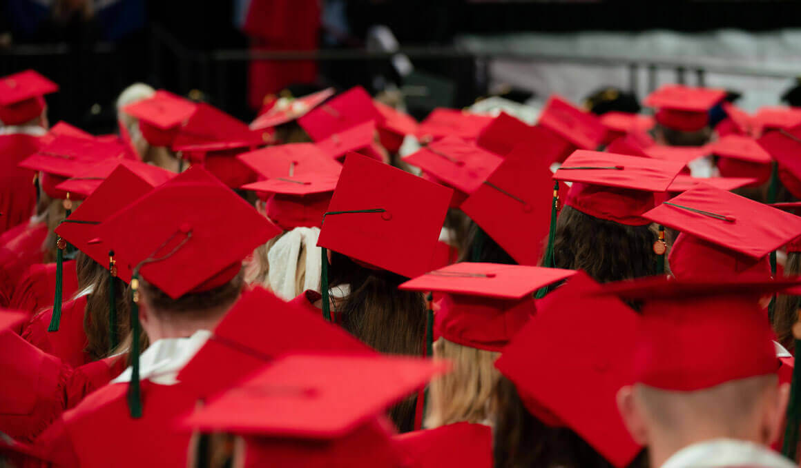 This photo shows an image of a group of USD students at commencement, with their backs turned to the camera. The focus of the photo is on their red graduation caps, that have a tassle hanging off to the left side.