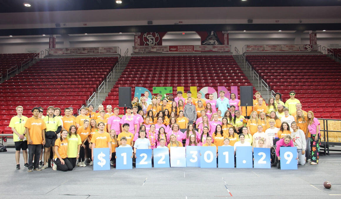 Participants of the 2026 Dakotathon sit together for a group photo in the Sanford Coyote Sports Center. They are wearing bright yellow, pink, and orange shirts and smiling at the camera. In front of them, there are blue boards that represent the total amount they raised for the 2026 Dakotathon.