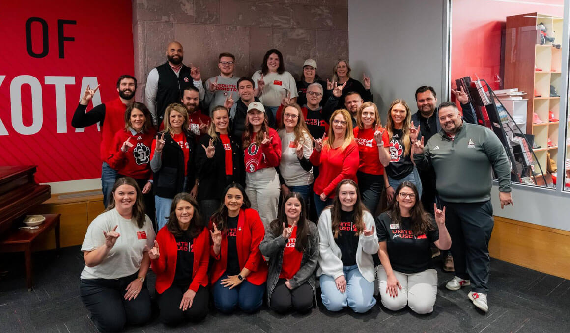 A large group of smiling people wearing University of South Dakota apparel poses indoors, many flashing the "Go Yotes" hand sign and wearing "Unite for USD" shirts.