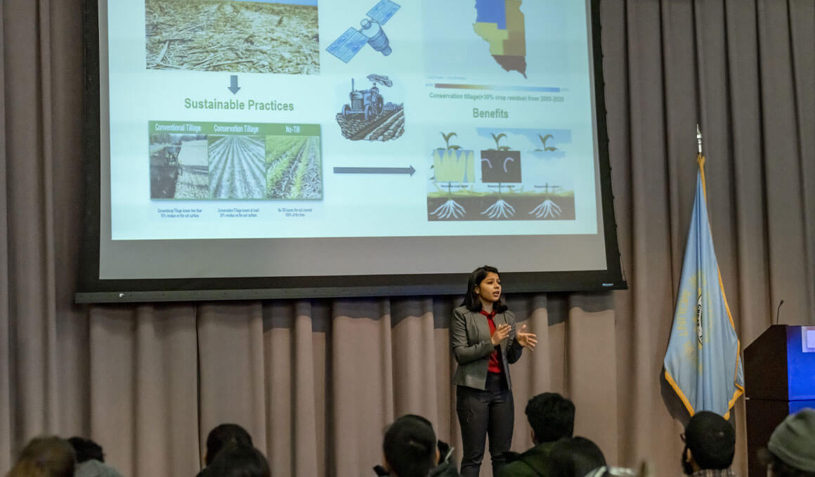 A female USD student stands on stage, with a PowerPoint on the screen behind her, which sits against a gray backdrop. She is presenting her research to a crowd. The state of South Dakota's flag is to her left, along with a podium.