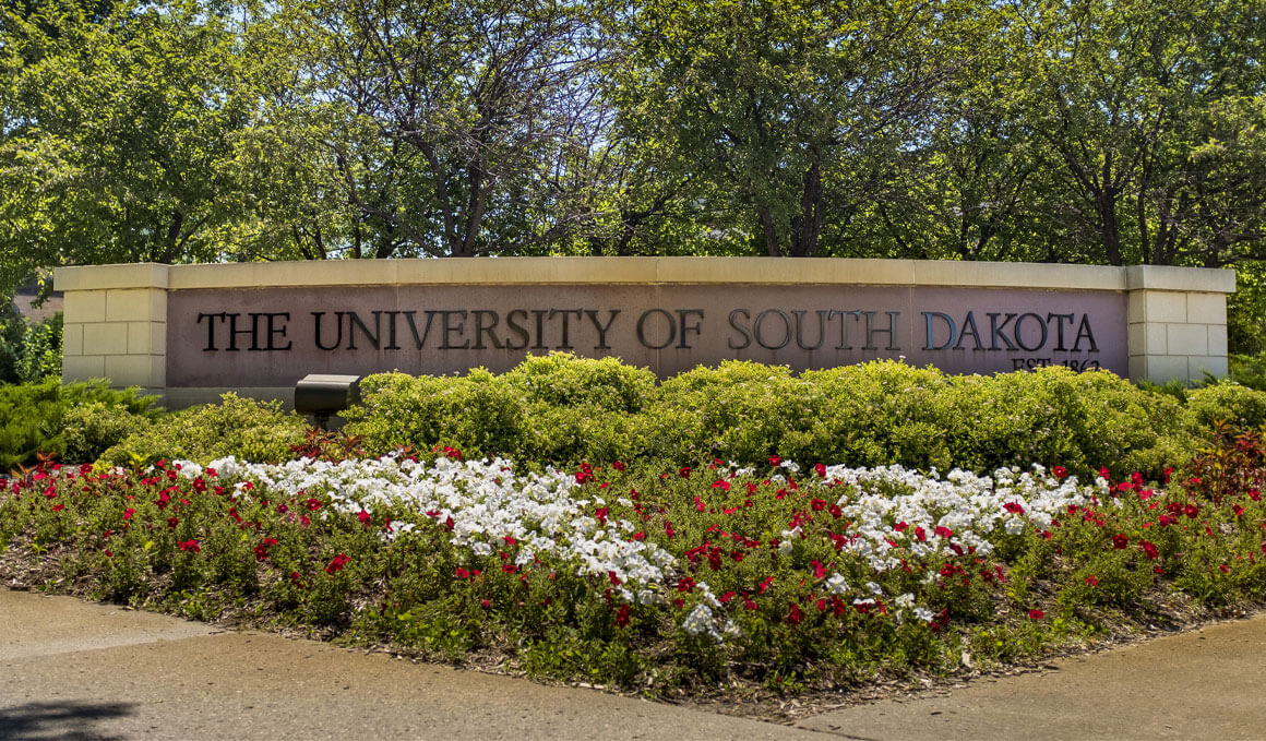 A stone and brick wall displays "The University of South Dakota." It is surrounded by spring flowers, green bushes and green trees.