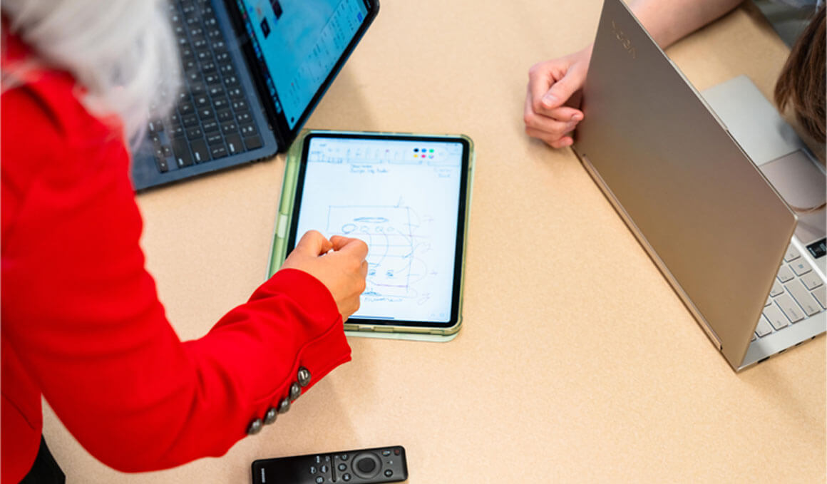 A USD professor works with students at a table. On the table there are laptops and an iPad, in which the professor is drawing on.