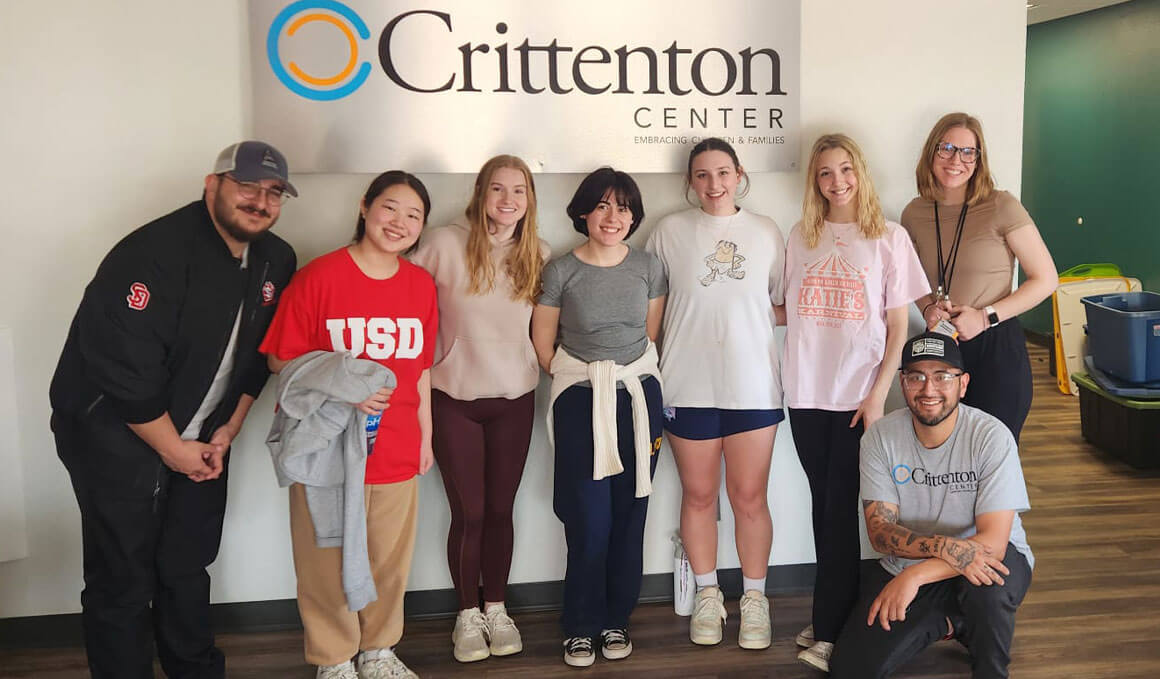A group of eight USD students pose together in front of a white wall featuring the Crittenton Center logo. The group is dressed in casual clothing; one student wears a red "USD" t-shirt. They are gathered in a bright, modern lobby area, volunteering their time.