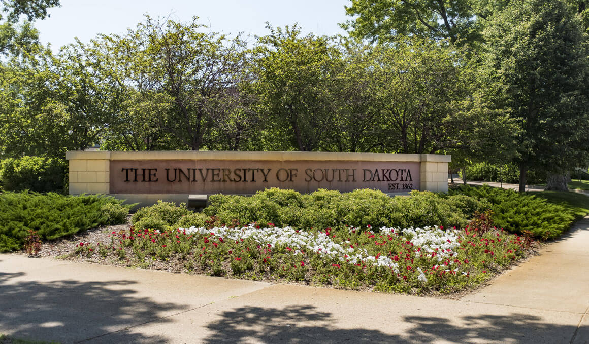 A brick sign that displays "The University of South Dakota" sits on the USD campus. It is surrounded by trees in the background and flowers and bushes in the foreground.