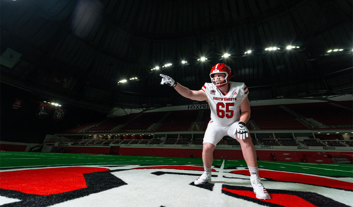 Caden Ellingson stands on the turf of the DakotaDome. He is dressed in his football gear, wearing his helmet and pointing.