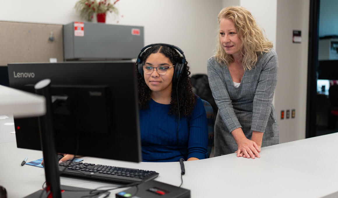 Angela Jackson, Ph.D., collaborates with a staff member in the Center for Digital Accessibility. The staff member is sitting in front of a computer and wearing black headphones.