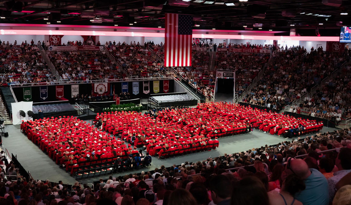 This photo shows a zoomed out shot of the USD commencement ceremony. In the middle is a sea of students, wearing their red regalia.