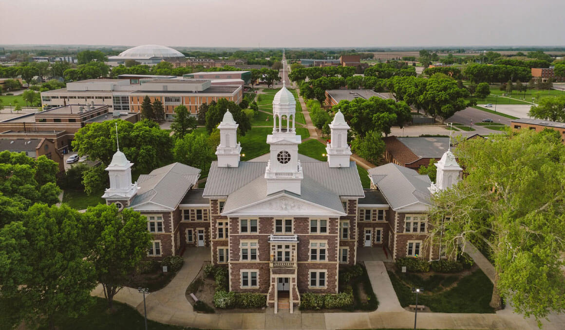 A drone image of the historic Old Main building, which sits on the USD campus on an overcast day. It is surrounded by bright green trees and other campus buildings.