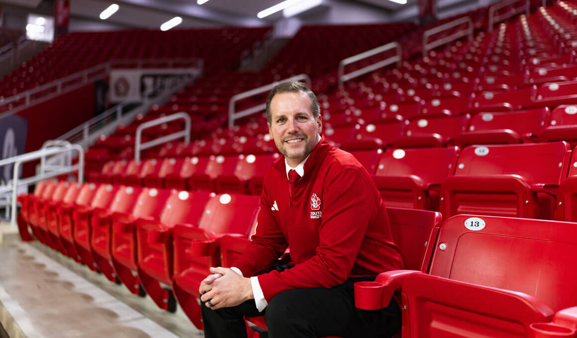 USD Athletic Director Jon Schemmel sits in the stands inside the DakotaDome. He is looking at the camera and smiling, slightly leaning forward with his hands clasped on his knees. He is wearing USD gear and is surrounded by bright red stadium chairs.