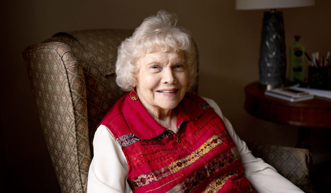 Nancy McCahren sits in a chair in her home while being interviewed for a story. She is wearing a long-sleeved white shirt with a red vest and matching earrings. She is looking at the camera and smiling.