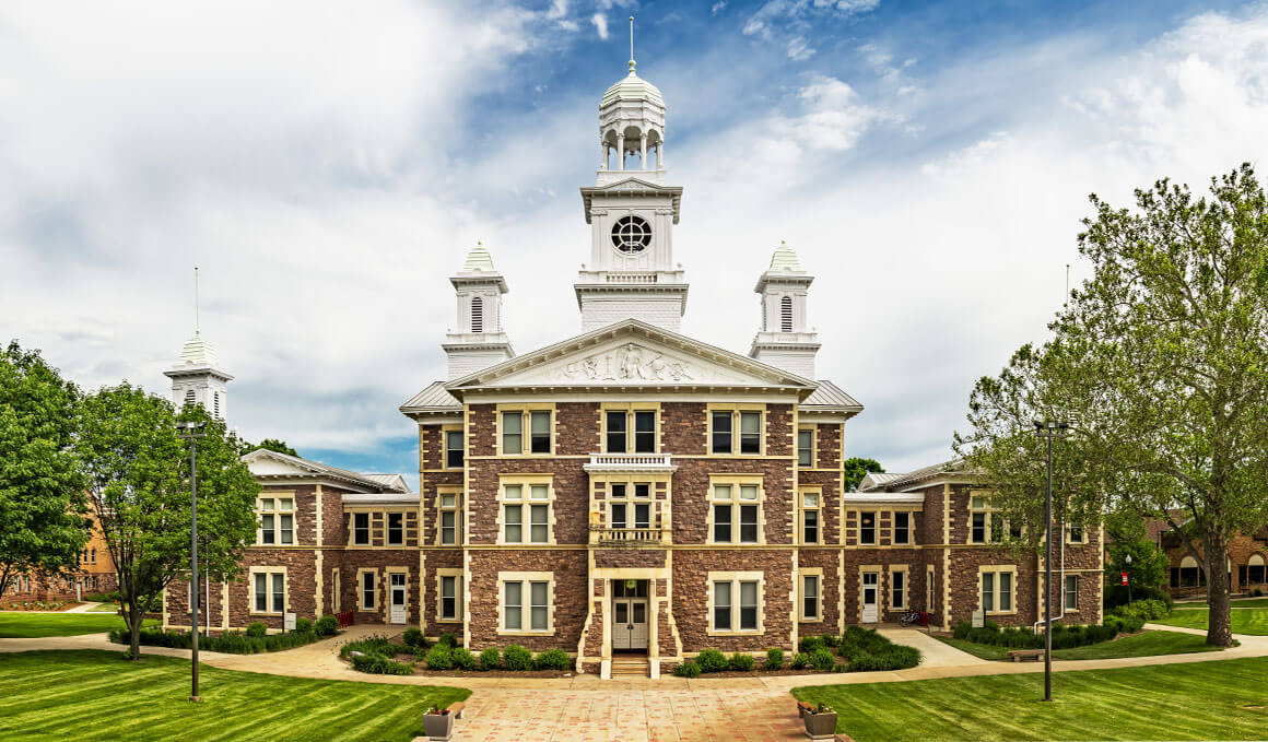 Old Main sits on the USD campus, surrounded by green grass and trees, with a cloudy blue sky in the background.