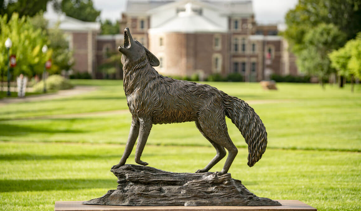 The Coyote Legacy statue sits on a sunny USD campus with Old Main in the background.