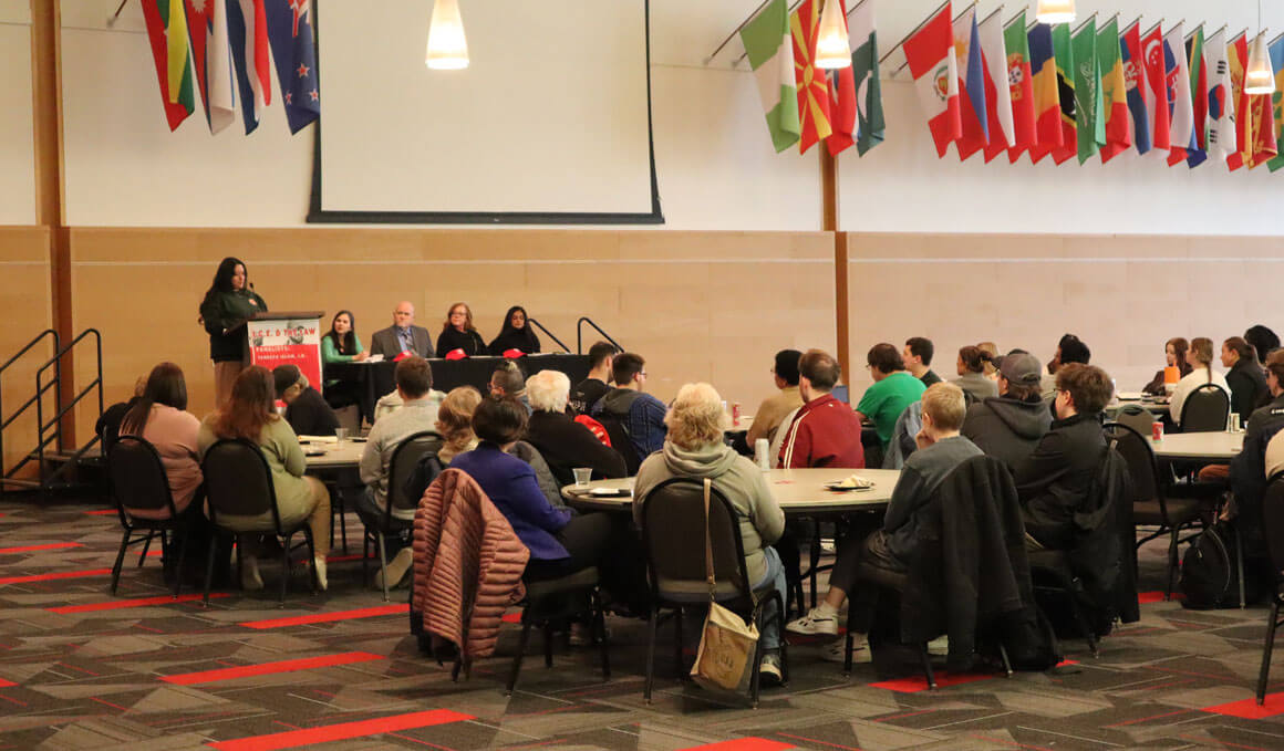 The Knudson School of Law student organizations hosts a panel. The audience is sitting in chairs at round tables, while looking towards a stage. The stage has five people on it, one of whom is standing at a podium speaking.