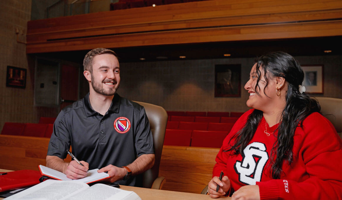 Two USD Knudson School of Law students, male and female, sit in the law school's court room. There are documents in front of them and they are looking at each other and smiling.