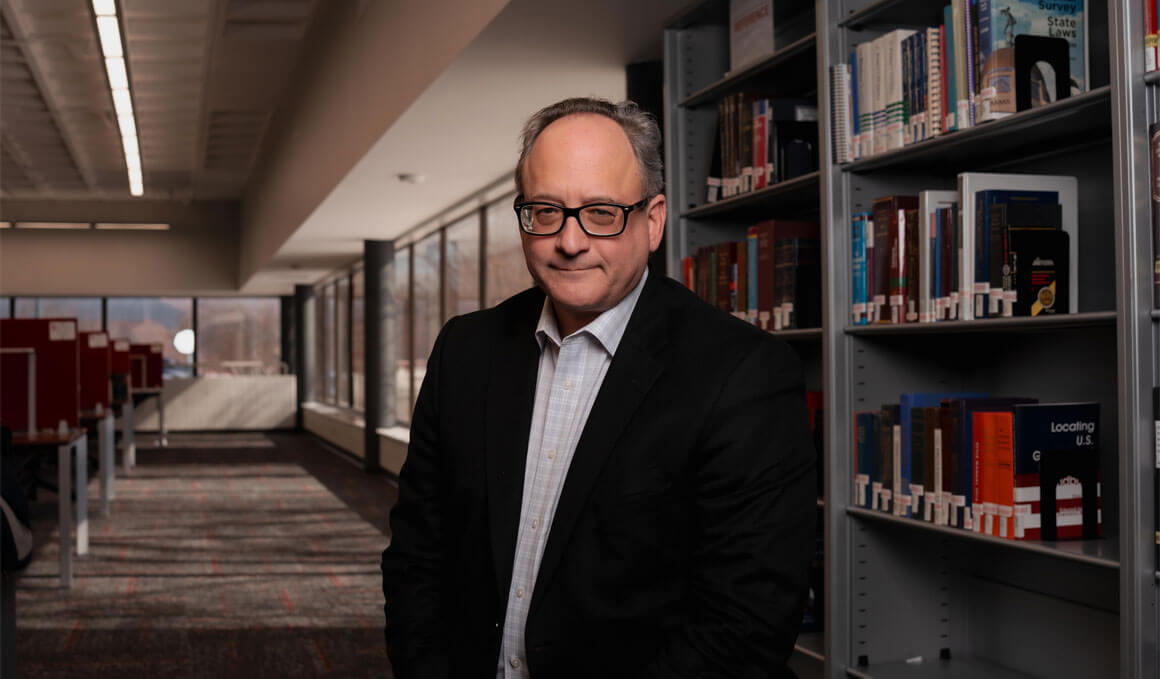 Tom Simmons stands in the McKusick Law Library, in front of a row of books and windows in the background. He is wearing glasses and a black blazer with a blue undershirt. He is smiling at the camera.