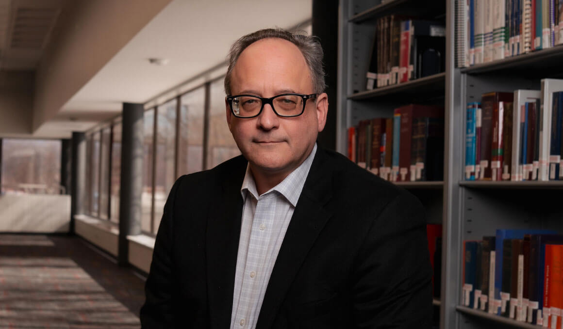 Tom Simmons,  J.D. ’98, stands in the law school's library. He is wearing a suit and glasses, and smiling at the camera.
