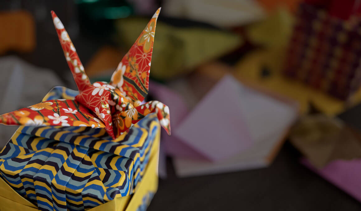 This image shows two paper origami figures. The first is a flowered, red, white and orange crane, sitting on top of a blue and yellow box.