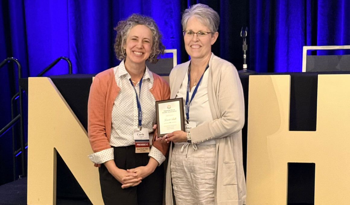 Janet Fulk, assistant director of the Frontier And Rural Medicine program at the USD Sanford School of Medicine, holds her plaque from the National Rural Health Association. She is standing with another woman and a bright blue curtain is behind them, along with the big letters "NH" in yellow cardboard.