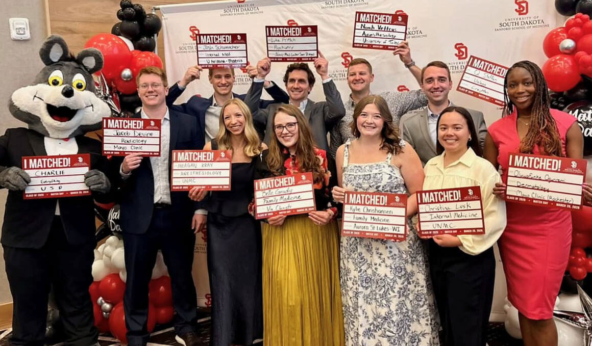 A group of USD Sanford School of Medicine students stand together, holding their match day signs and smiling. Charlie Coyote is standing next to them and they are all standing in front of a USD branded backdrop.