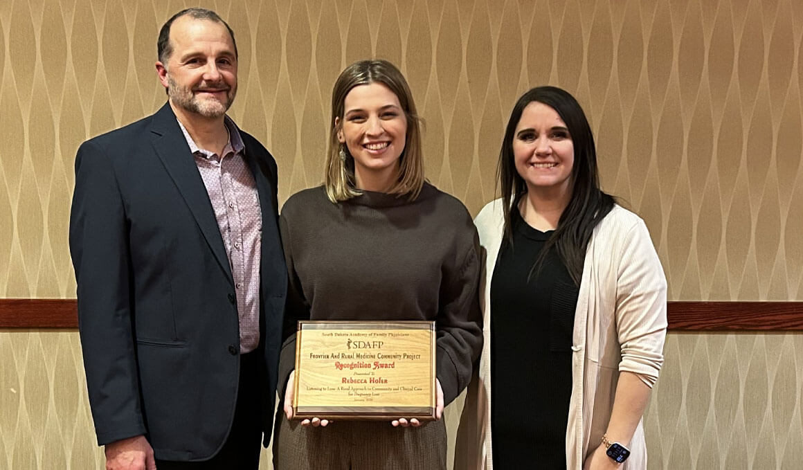 Rebecca Hofer stands against a wall, holding an award plaque. There is a man on her right side and a woman on her left. All are smiling at the camera. 