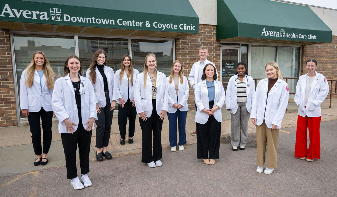 Eleven University of South Dakota Sanford School of Medicine students stand outside of the Coyote Clinic. They are dressed in their white lab coats and smiling at the camera.