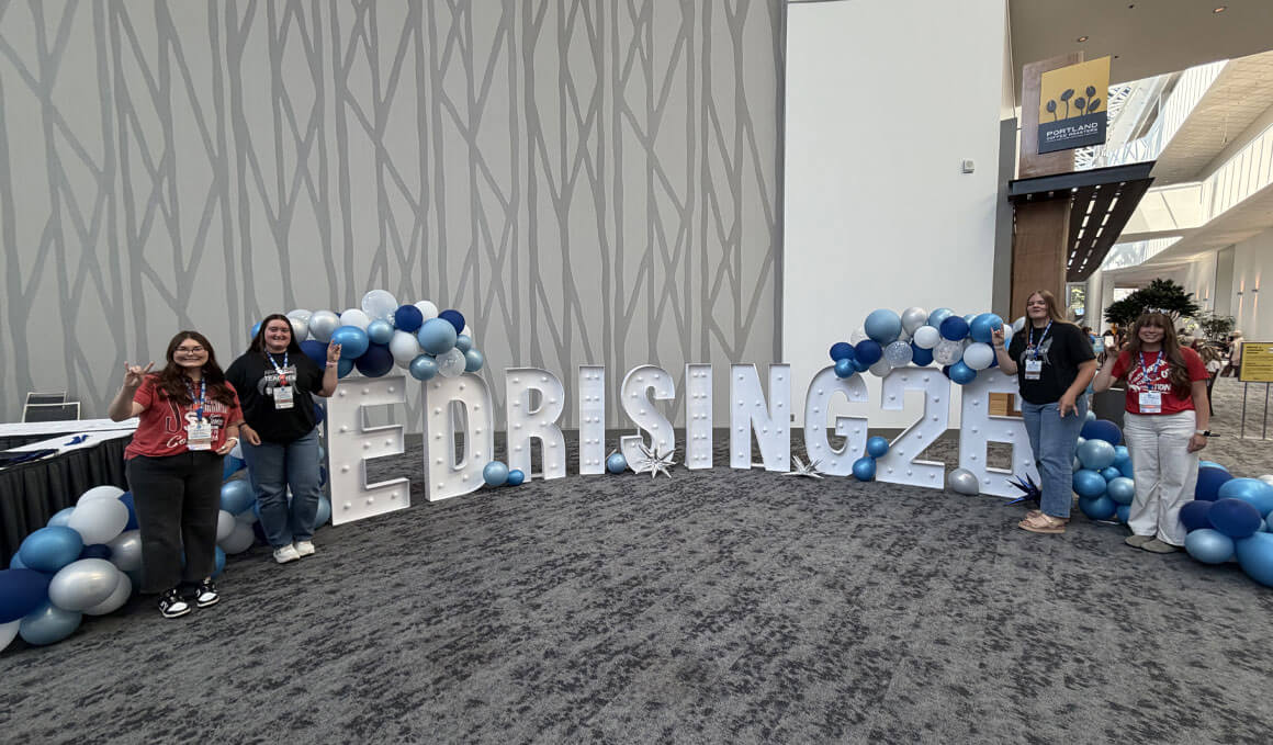 Students stand in a large conference room, around big, individual letters that read, "ED RISING 26." There are blue and white balloons on both sides of the sign.
