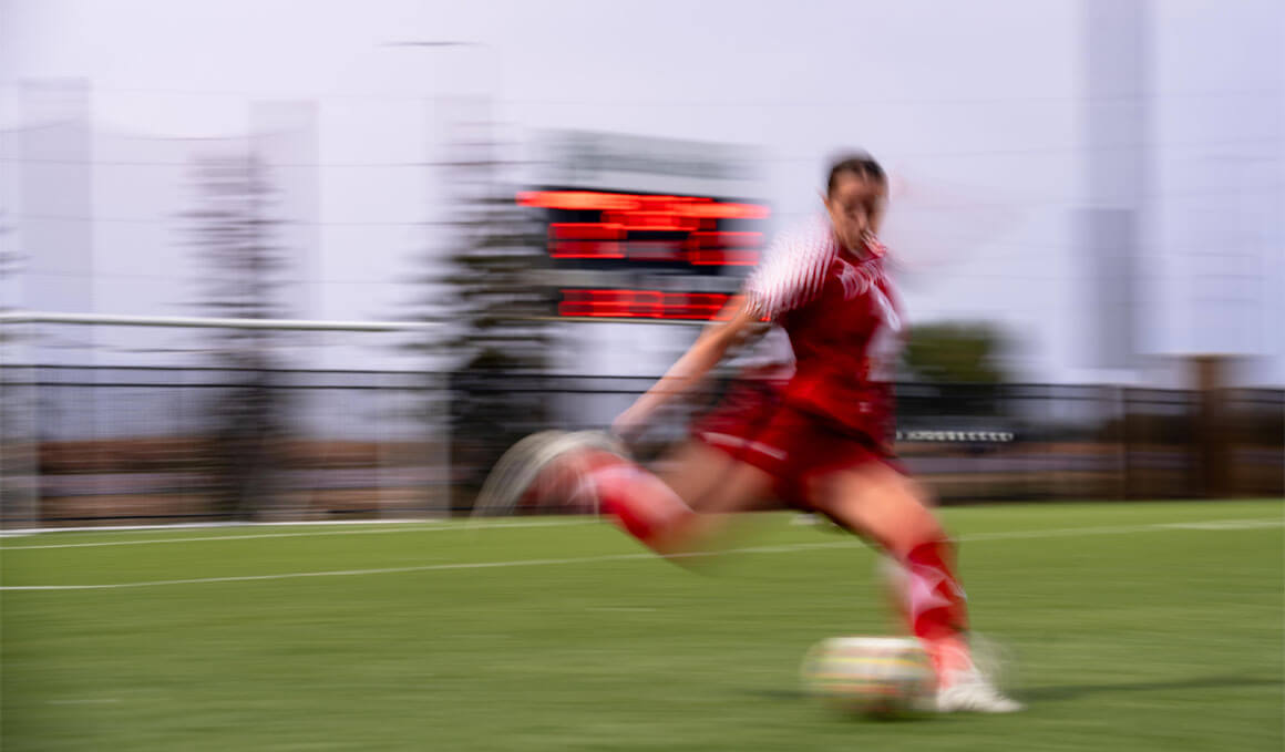A motion-blurred action shot of a female soccer player in a red uniform kicking a soccer ball on a green turf field. In the background, a scoreboard and the silhouette of trees are visible under an overcast sky, capturing the high-speed movement of athletic performance.