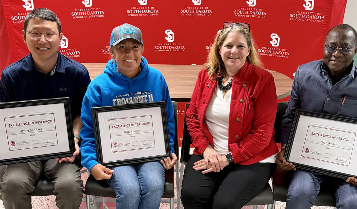 USD School of Education faculty members sit in chairs, holding their plaques for the Excellence in Teaching Awards. They are sitting with Dean Amy Schweinle, Ph.D., and are seated in front of a red, branded USD backdrop.