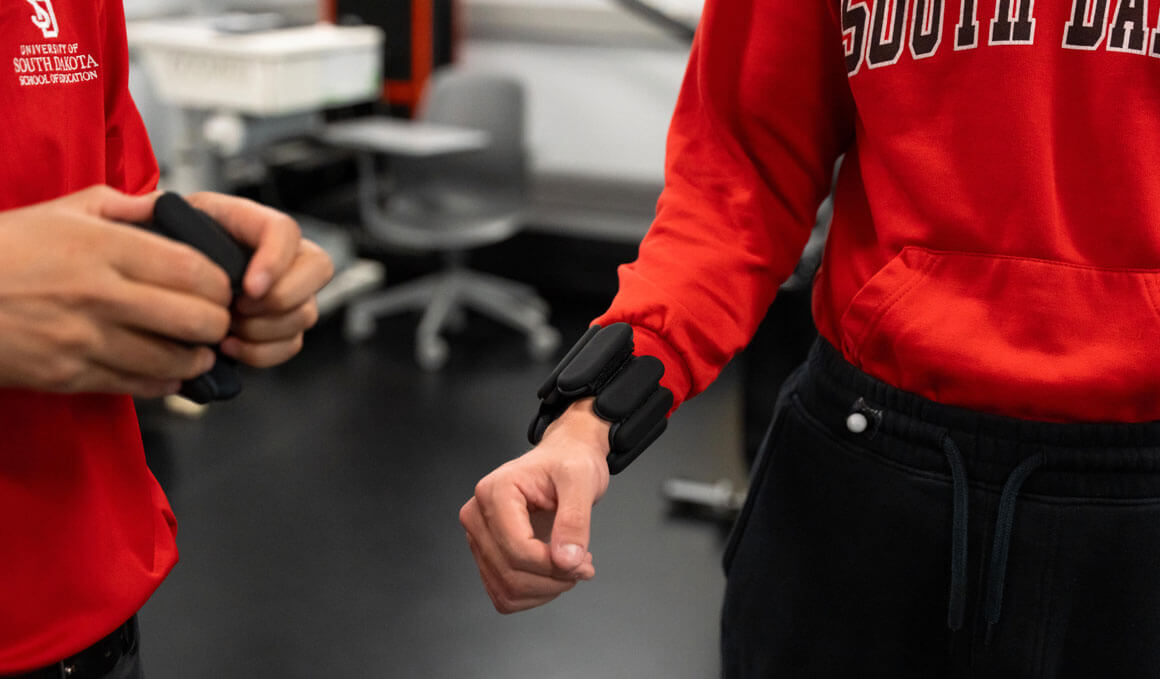 Two students (wearing red USD sweatshirts) adjust black wearable weights that are strapped to their wrists in a research lab setting.