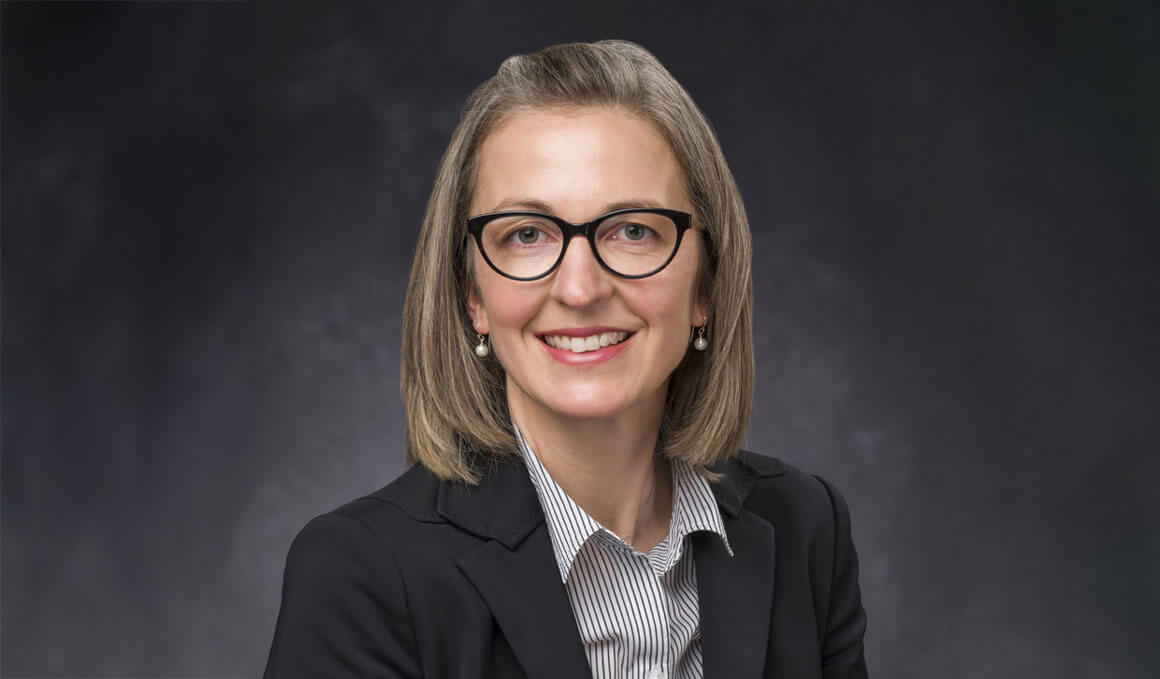 Becca Jordre poses for her professional headshot. She wearing glasses and smiling, while wearing a dark blazer against a neutral studio background.