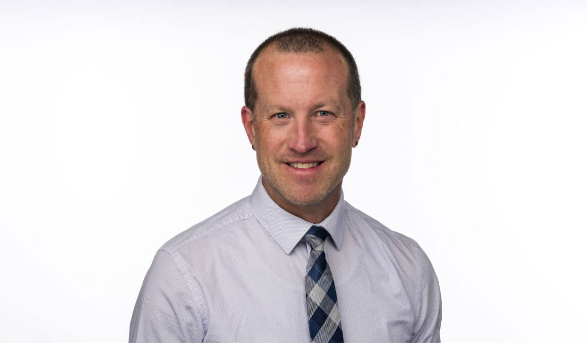 Kelly Bass, DSW, sits and poses for his professional photo. He is wearing a long-sleeved button up shirt with a black checkered tie. He is smiling at the camera.