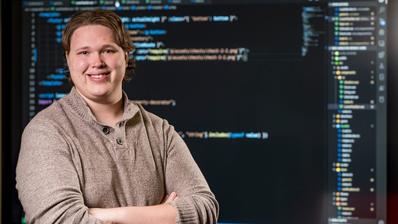 Person smiles in front of screen with computer code on it.