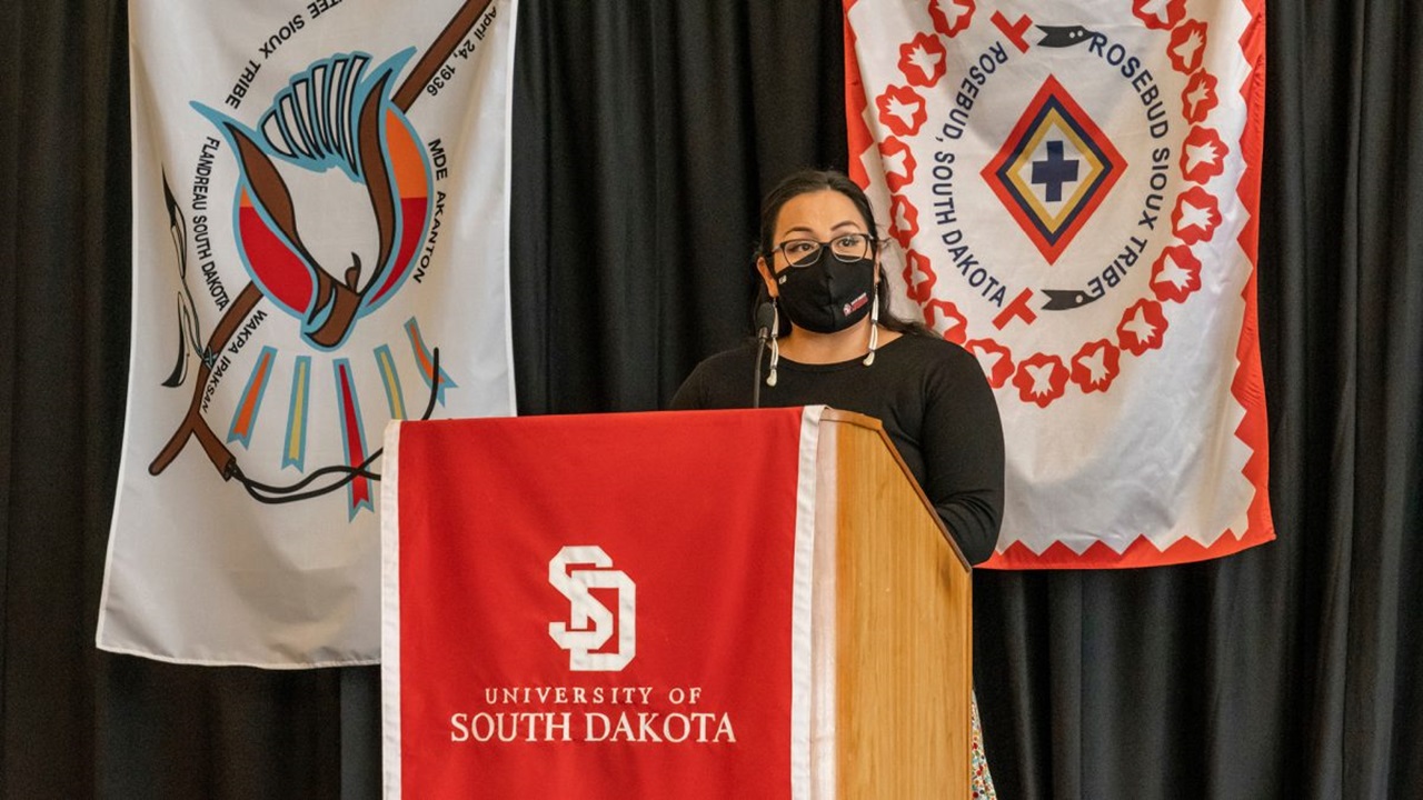 A woman stands behind a USD podium and gives a speech.