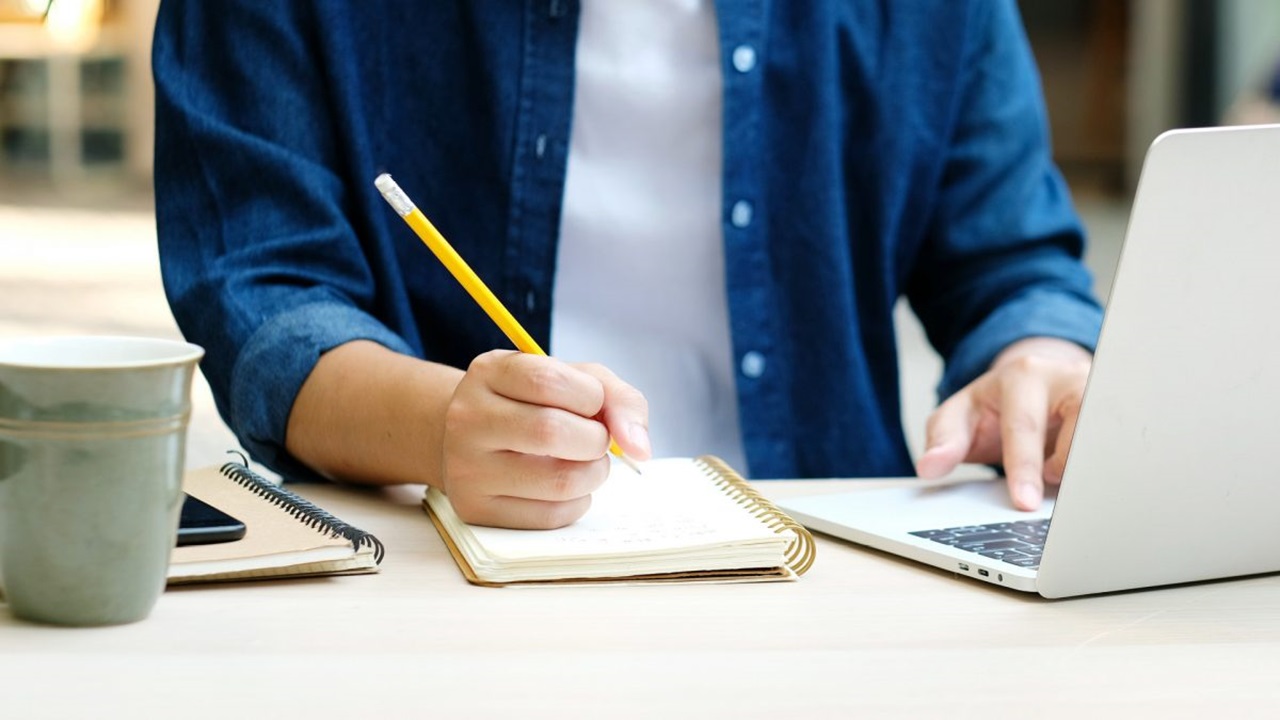 Person takes notes next to an open laptop.