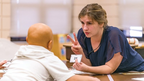 A woman holds up the number two on her hands and uses flashcards to teach a student.