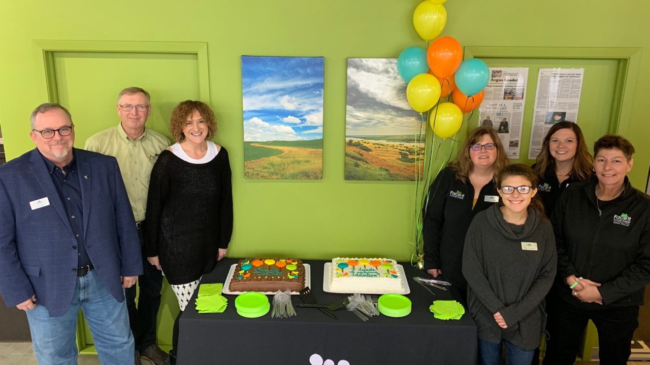 A group of people poses around a table with decorated cakes and balloons.