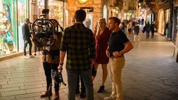 A film crew discusses in a low-lit area with shops. They hold a camera and plan their next shot.