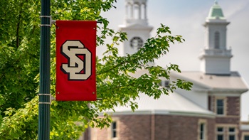 A close up photo of a red USD banner on campus. Old Main is in the background.