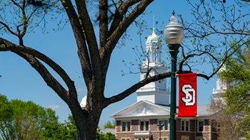 A photo of a USD banner on campus in the spring. Old Main is in the background. 
