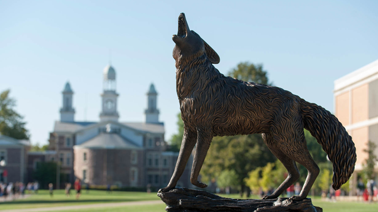 A howling coyote statue with Old Main in the background. The grass is green and students walk on the sidewalk behind the statue.