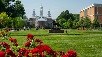 Red flowers are in focus, with bright green grass in the background. The image shows the Muenster University Center lawn on USD's campus, with the Legacy statue and Old Main both visible.