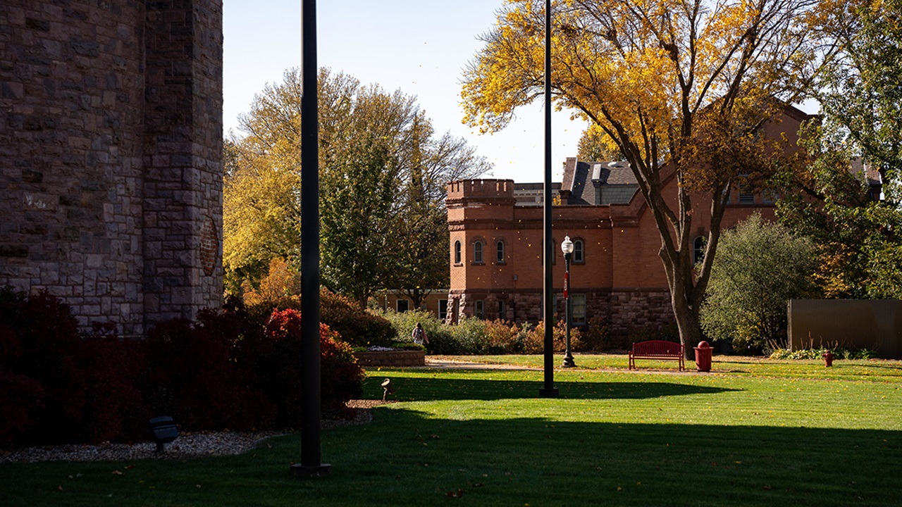 Dramatic lighting cuts across a greenway with brick buildings in the fore and background, with trees in fall foliage.